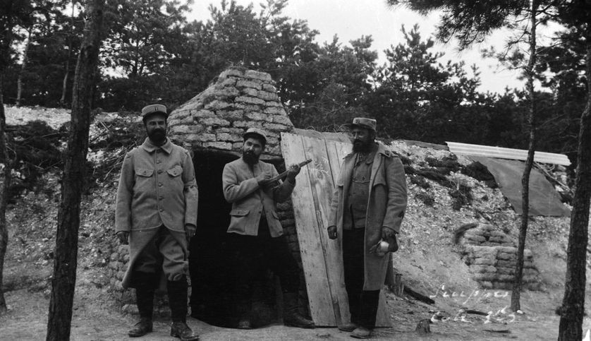 A l'entrée d'un abri, l'un des trois soldats joue sur un instrument de musique bricolé avec du bois. Trigny, Marne, 1917.