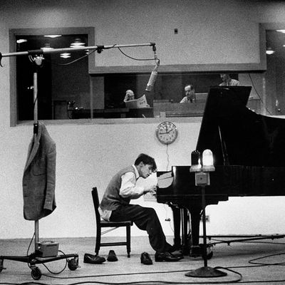 Glenn Gould playing concert grand piano as he records Bach in recording studio while engineers & dir. following score look on fr. glassed in booth as boomed mike doubles as his coatrack.