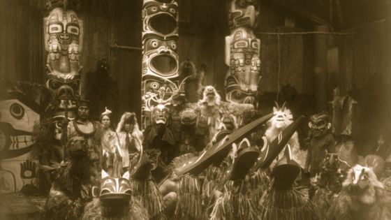 États-Unis, vers 1914: au cours de la cérémonie d'hiver, les danseurs Kwakiutl portent des masques et des costumes. Ils posent devant Trois mâts totémiques. Le chef à l'extrême gauche tient un bâton d'orateur. ©Getty - Photo de Buyenlarge / Getty Images