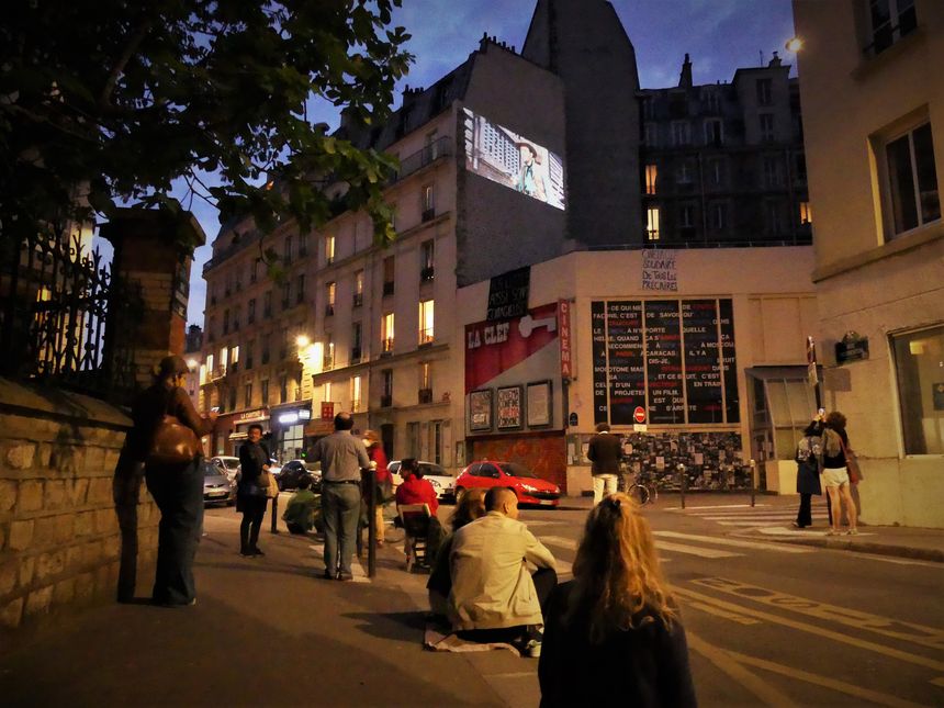 Des spectateurs sont installés sur le trottoir pour admirer cette projection en plein air.
