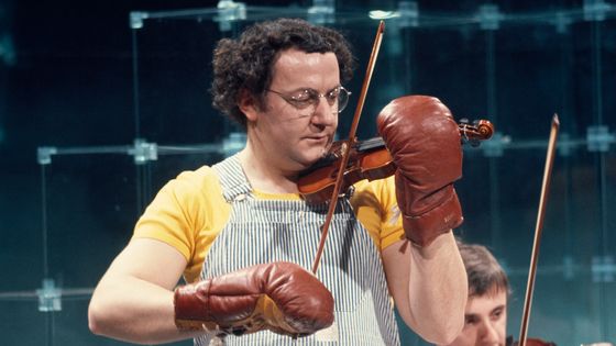 Portrait de l’humoriste et comédien Coluche jouant du violon avec des gants de boxe dans l'émission "Numéro Un". ©AFP - JACQUES CHEVRY / Ina