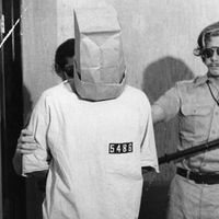 1971, un prisonnier forcé de porter un sac sur la tête, à côté le gardien John Loftus durant l'expérience de l'emprisonnement à l'université de Stanford